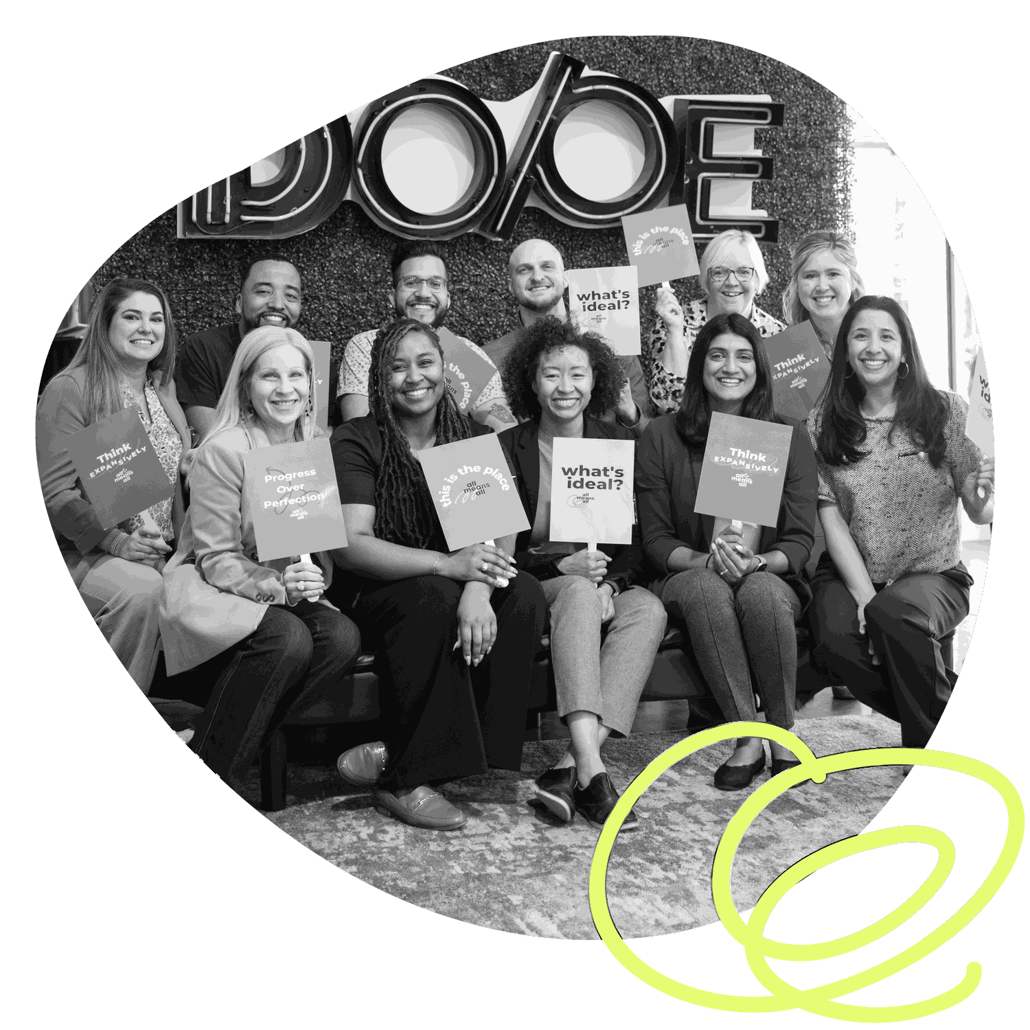 The All Means All team together at a convening, seated and standing in front of a greenery wall, holding cards that read 'what's ideal?', 'this is the place', 'think expansively' and 'progress over perfection.'